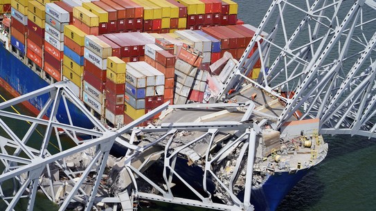 Wreckage of a collapsed highway bridge lies across the deck of a cargo ship.