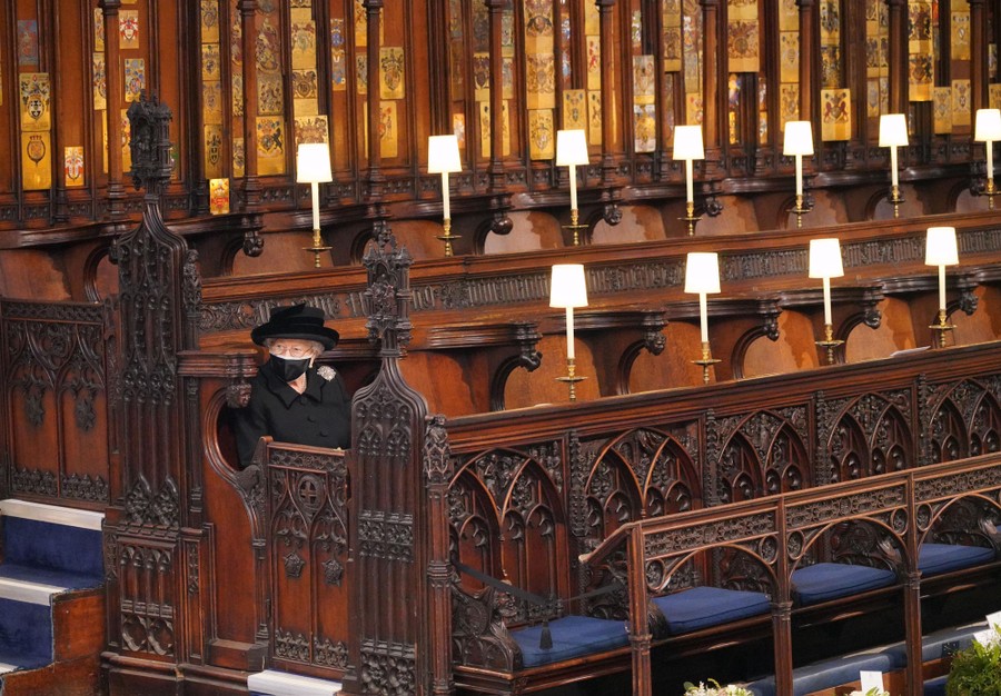 The Queen of England sits alone in an ornate chapel, dressed in black, wearing a face covering.