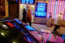 The neon lights of an adult-video store are reflected on a car.