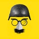 A military helmet floats above disguise glasses with fake nose and mustache. One lens of the glasses is shattered.