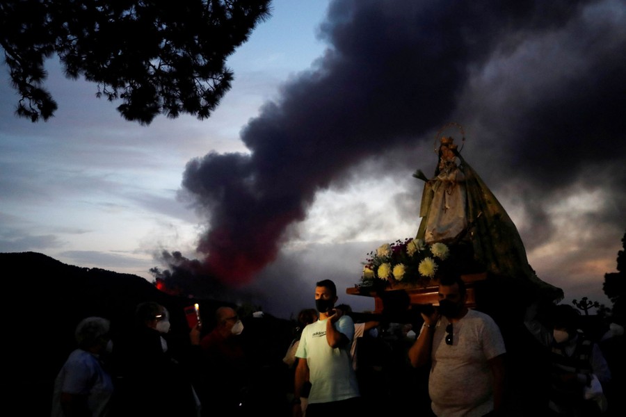People carry a small statue of the Virgin Mary outside as an eruption is seen in the background.