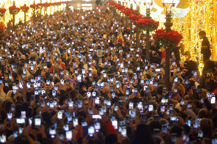 Hundreds of people in a crowd beneath holiday lights hold up their cellphones to take photos of the scene.