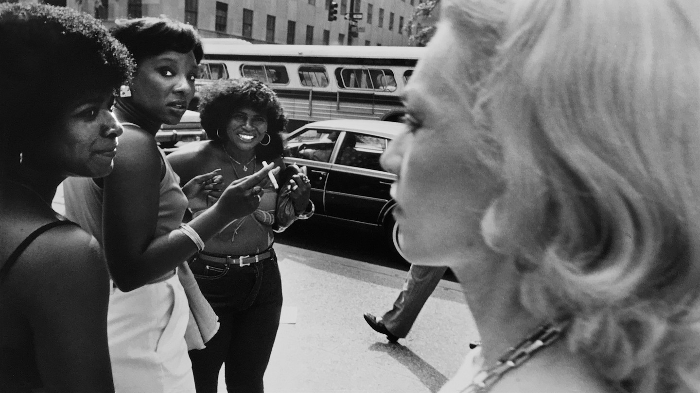 On the left side of frame, three Black women stand together on the street; on the right, a blond woman walks by with a serious face, and a man's leg is in the background
