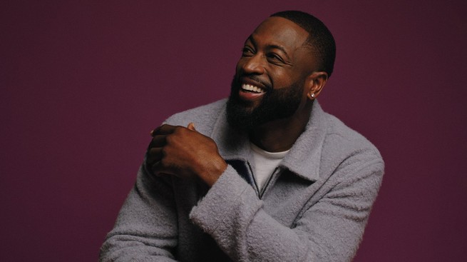 photo of Dwyane Wade smiling and sitting with arms crossed, wearing gray collared jacket, against purple background