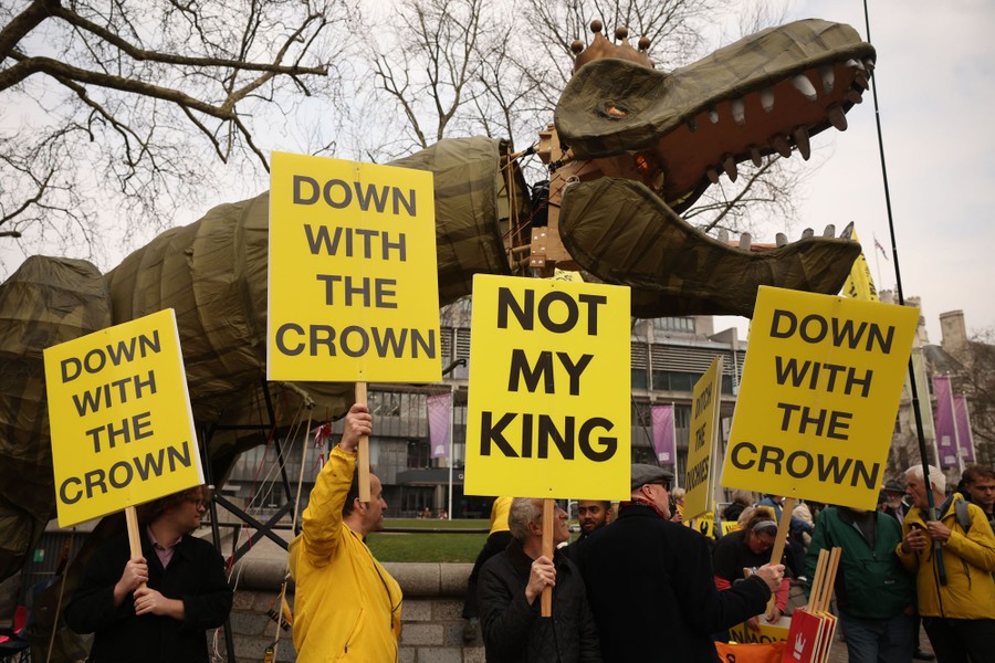 Anti-royalist protesters hold signs next to a dinosaur puppet wearing a crown, holding signs that read "Down with the crown."