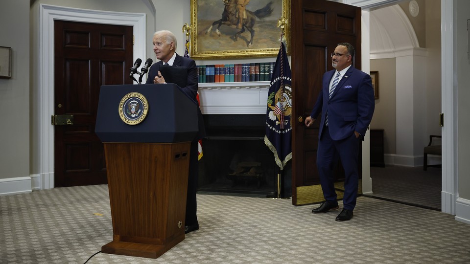 Joe Biden gestures at a White House podium, Miguel Cardona stands by the open door