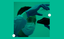 A man wearing glasses, a surgical mask, and rubber gloves examines laboratory samples in small vials.