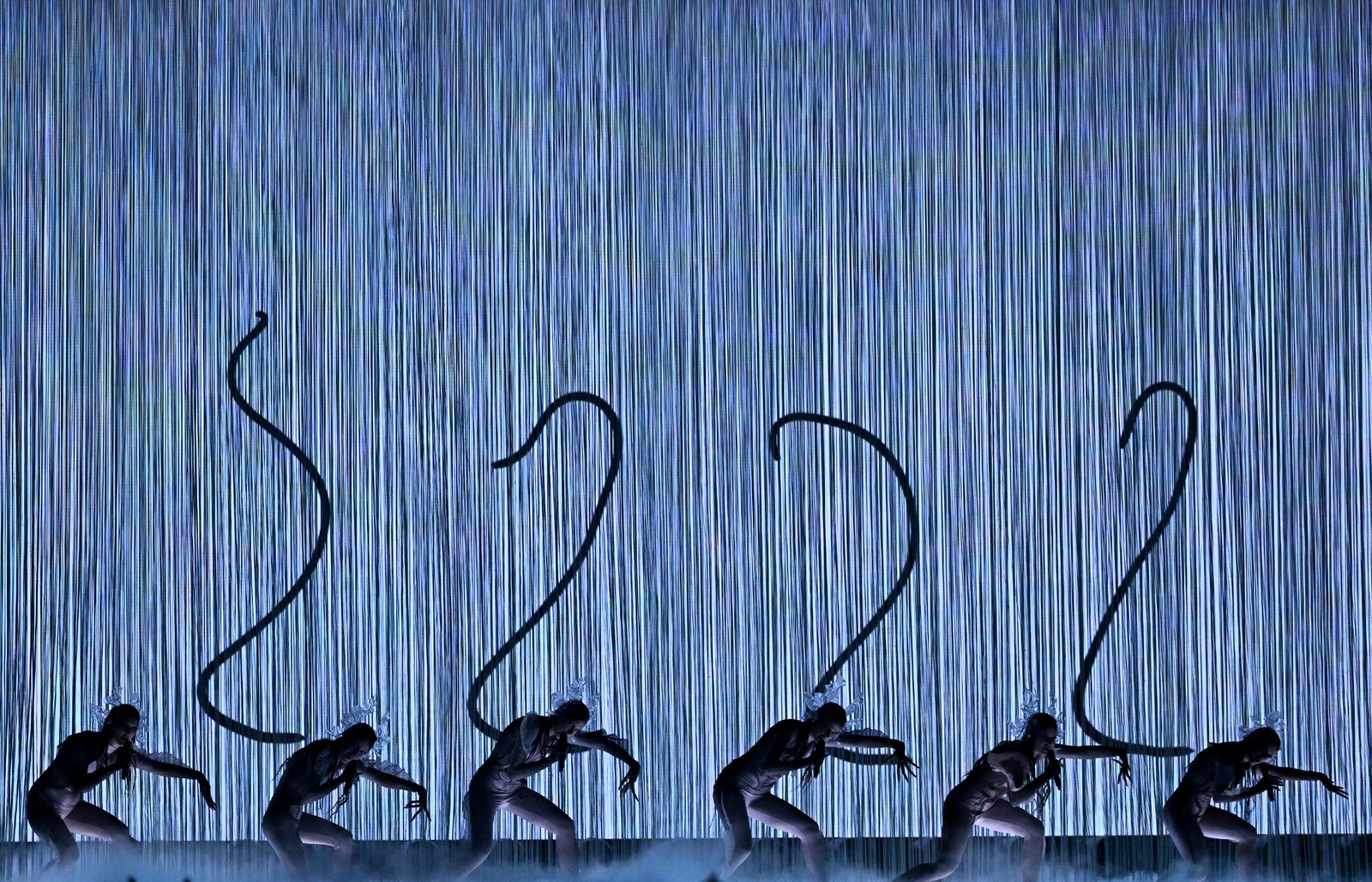 Six performers pose in front of a video projection that makes it appear as if they have very long tails.