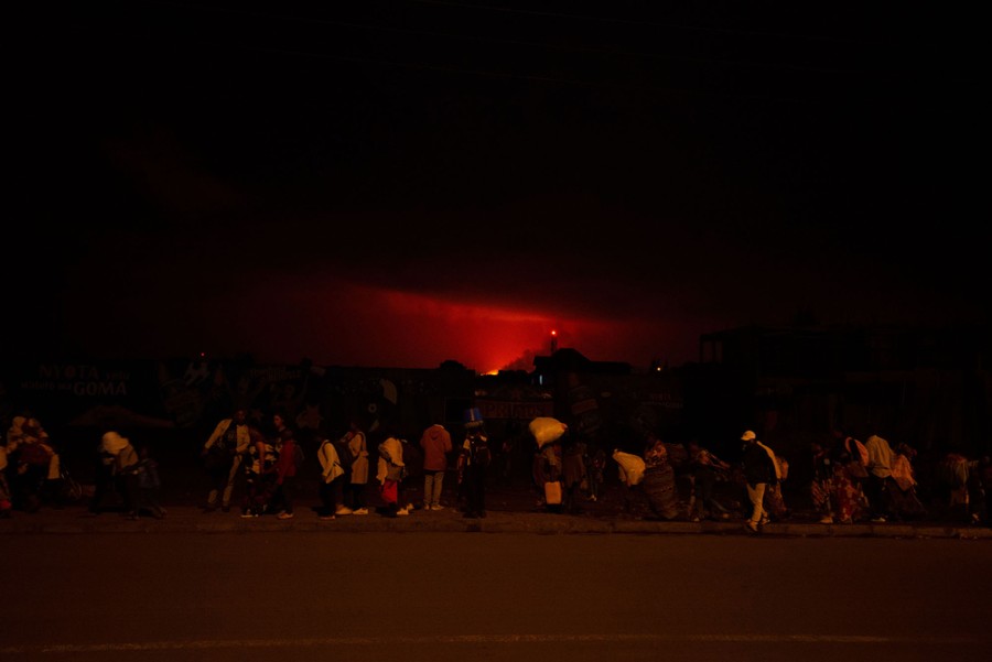 A line of people walk along a street, carrying belongings, at night.