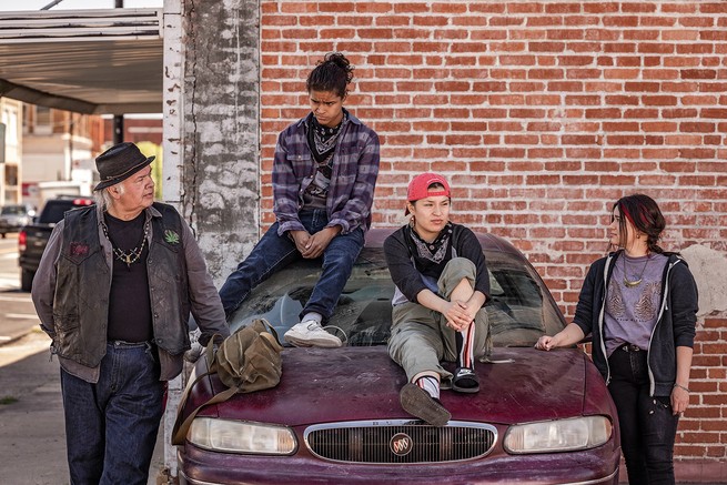 Photo of older man in hat and jacket leaning on the hood of a burgundy car with two younger people sitting on roof and hood and one on other side, in front of brick wall.