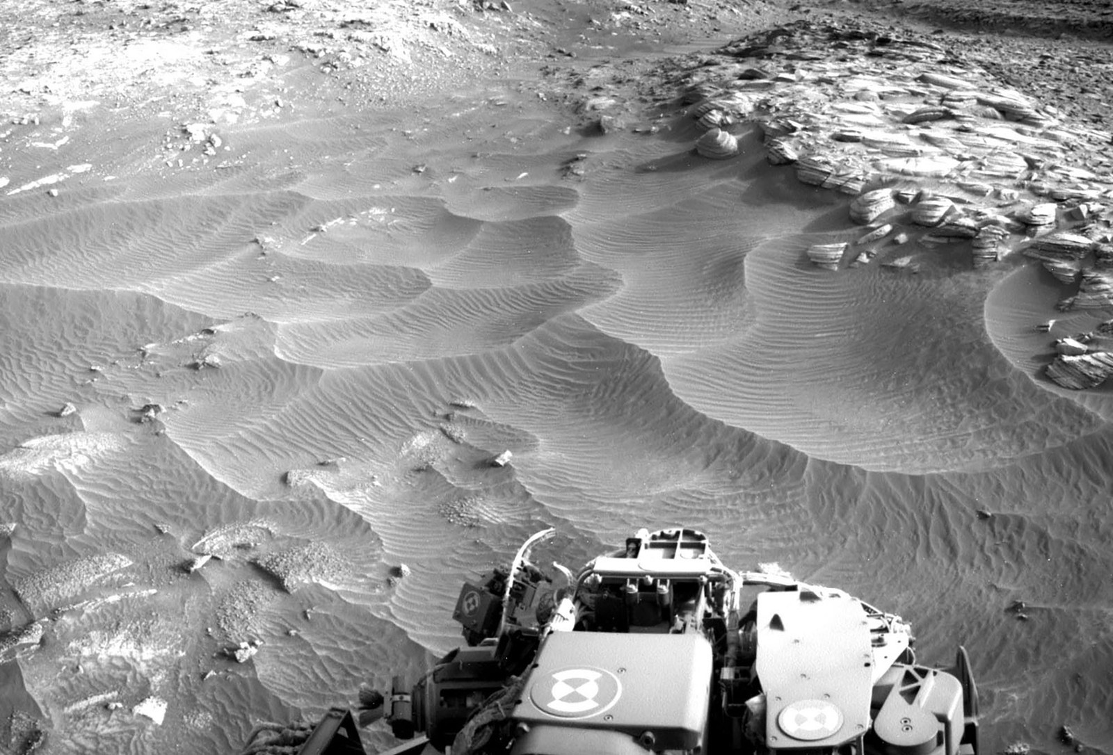 Part of a Mars rover, seen beside rippling sand dunes and a rocky outcrop