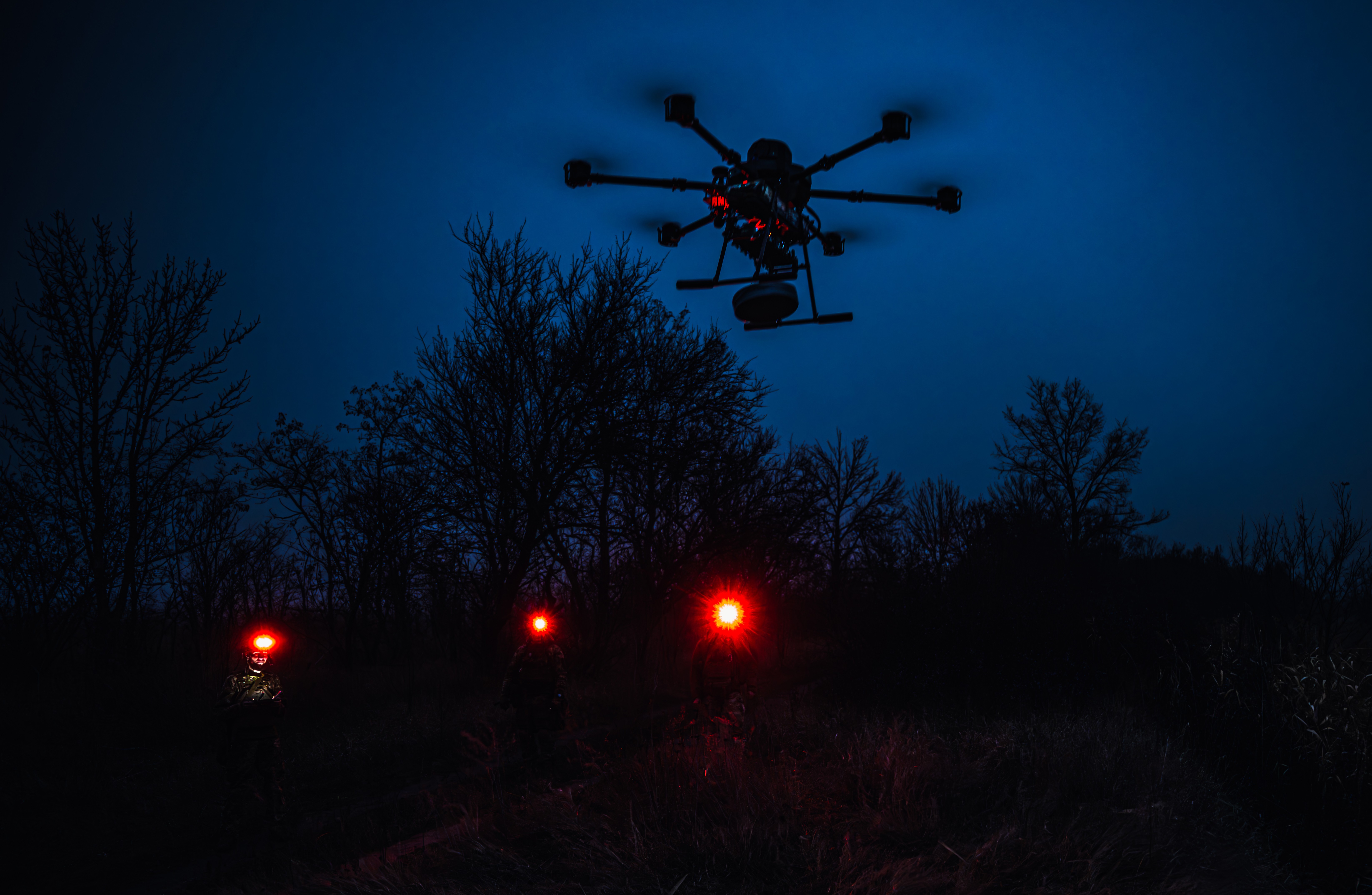 Several soldiers with red headlamps stand near a large hovering drone at night.