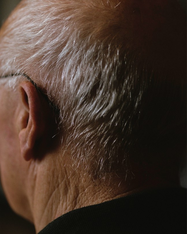 A man's head from behind, showing his ear and gray hair and fading into black