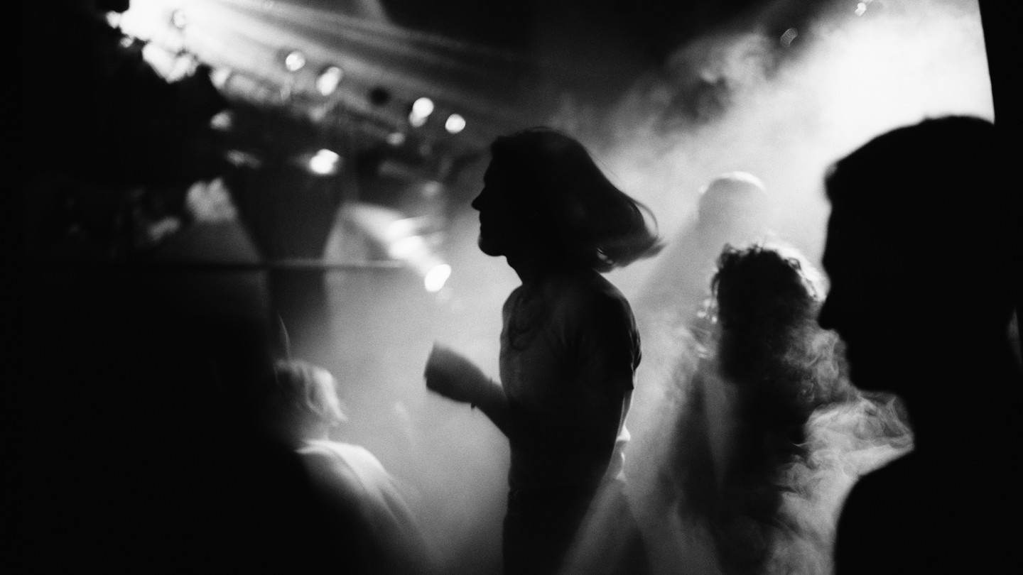 A black-and-white photograph of a person with mid-length hair, covered in shadow, dancing in a smoky room with other people