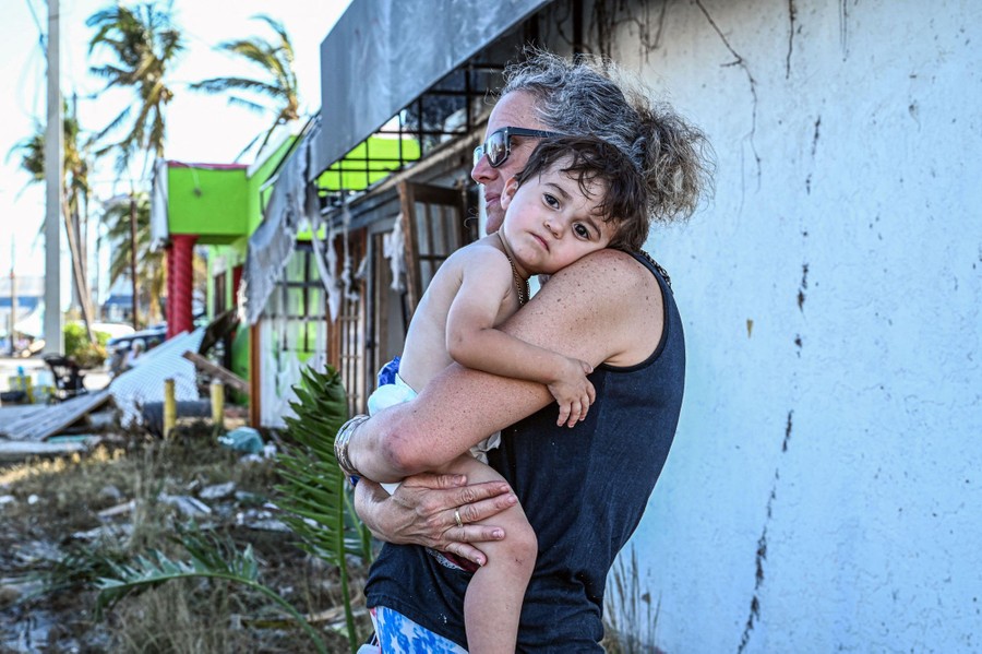 A woman hold a small child in her arms outside a building, with storm damage visible in the background.