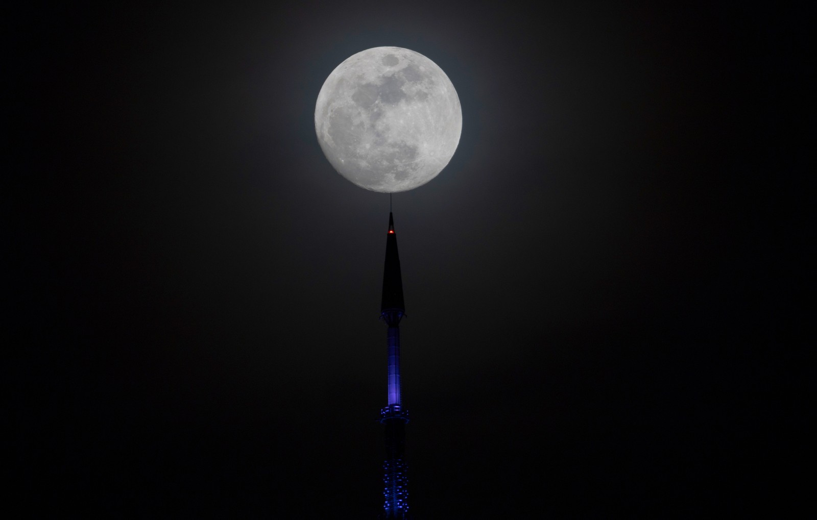 A full moon rises behind the antenna of a skyscraper.