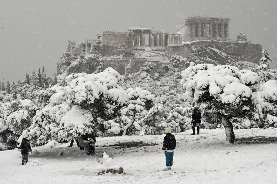 People play in the snow, with ancient Greek ruins visible in the background.