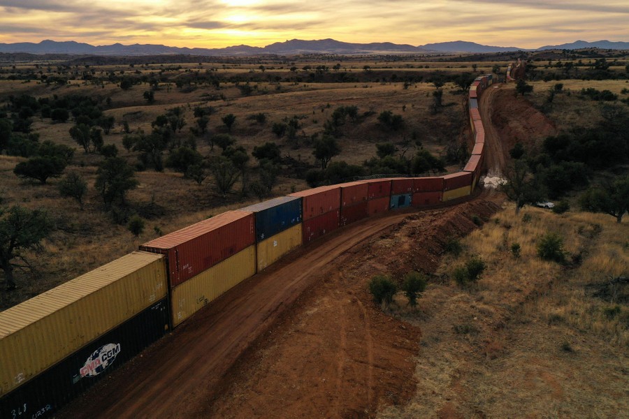 A long line of shipping containers, stacked two high, runs parallel to a dirt road through a semi-arid stretch of wilderness.