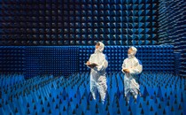 Two technicians dressed in white protective gear stand inside a test room covered in foam cones.