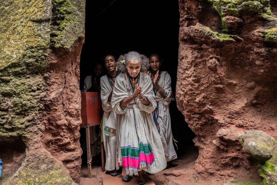 Several young women sing and walk through a stone doorway.