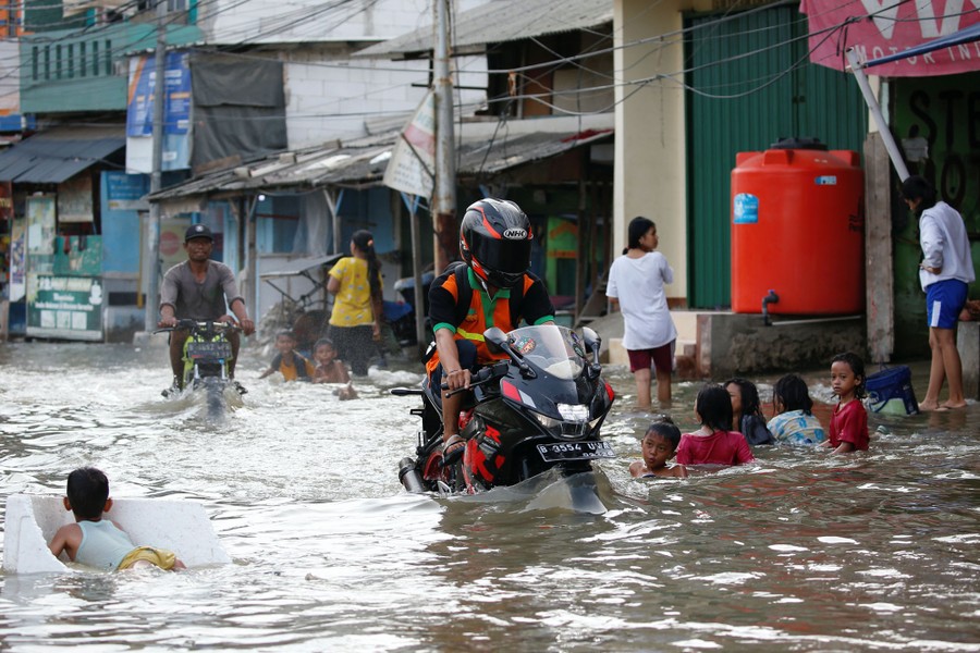 People walk, swim, and drive motorbikes through a flooded street.