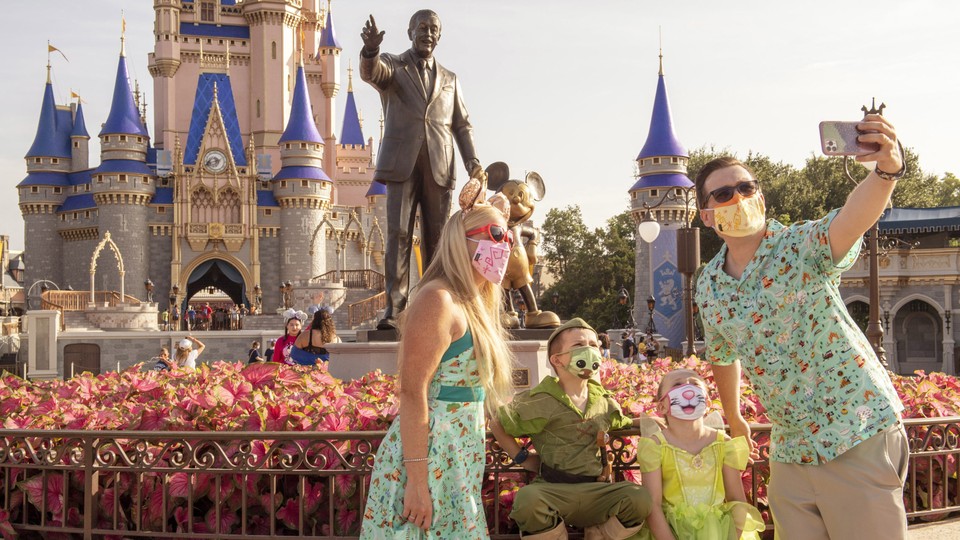 A family wearing masks takes a selfie in front of the statue of Walt Disney and Mickey Mouse at Disney World.