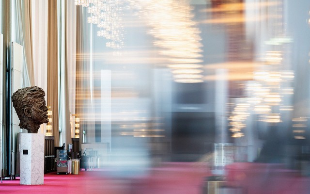 A blurred color photo of the Grand Foyer of the Kennedy Center, showing a bronze bust of John F. Kennedy in focus.