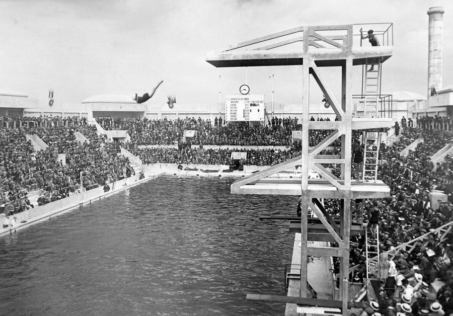 A person makes a high dive into a pool in an aquatics stadium filled with many spectators.