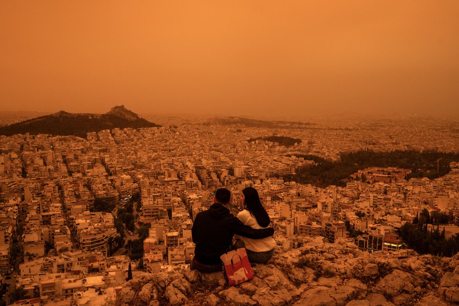 A couple sits on a hillside overlooking buildings in Greece, beneath a hazy orange sky.