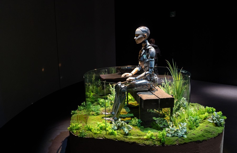 A robotic figure, displayed in an exhibit, seated on a bench surrounded by plants.
