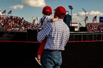 An adult man holds a child at a Trump rally. Both are wearing red hats.