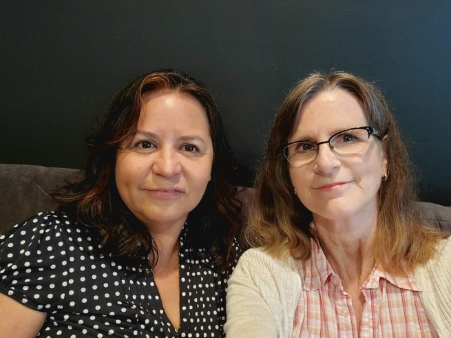 Two women sitting side by side on a couch, smiling into a camera. The one on the left is middle-aged, Latina, with dark brown hair, wearing a polka dot shirt. The one on the right is older, white, with greying brown hair, wearing glasses, a checkered shirt, and a cardigan.