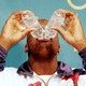 A man drinks from two plastic water bottles simultaneously. Both bottles are tipped over his mouth, blocking the upper half of his face.