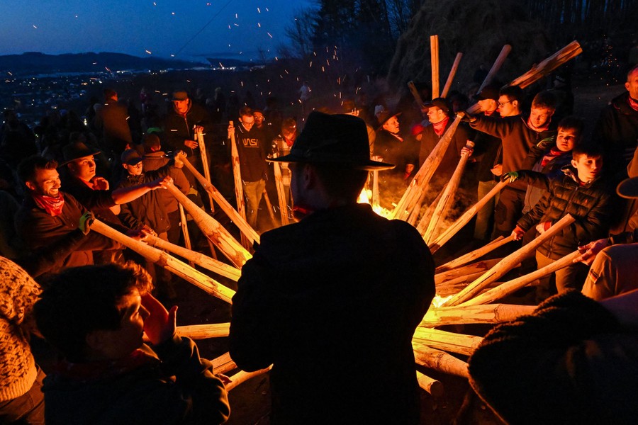 A small crowd of people gather in a circle around a fire, each placing a long wooden torch into the flames.