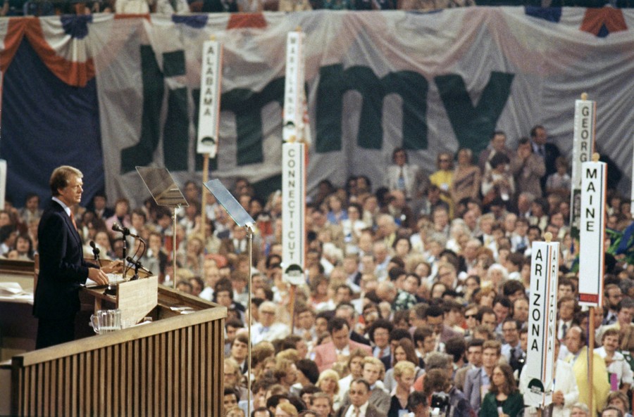 Jimmy Carter stands on a stage, speaking to a large crowd of delegates at a political convention.