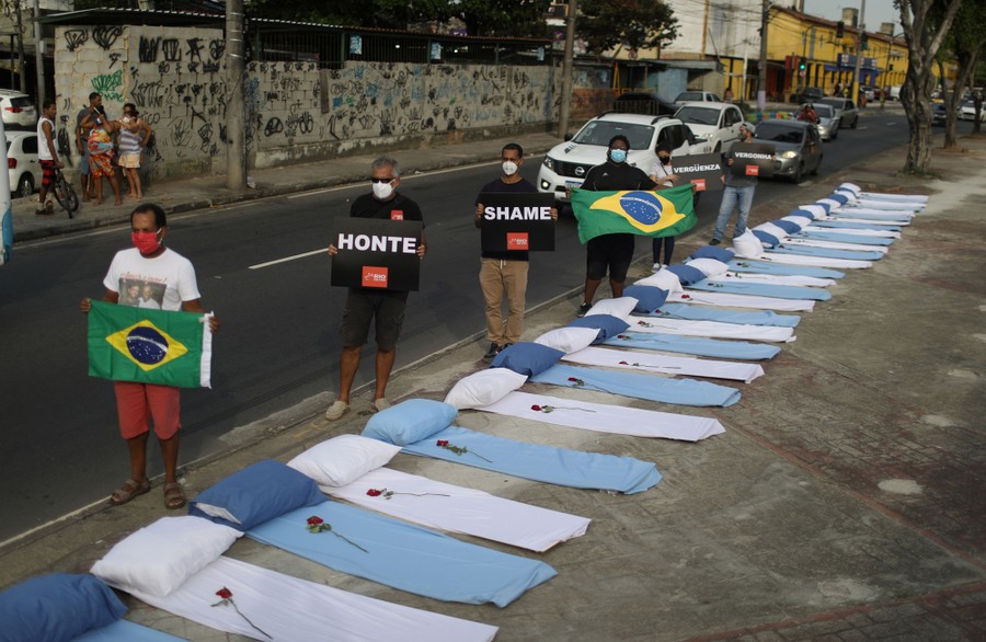Six protesters stand along a road beside dozens of pillows and sheets laid out like beds, holding Brazilian flags and signs that say "Honte" and "Shame."