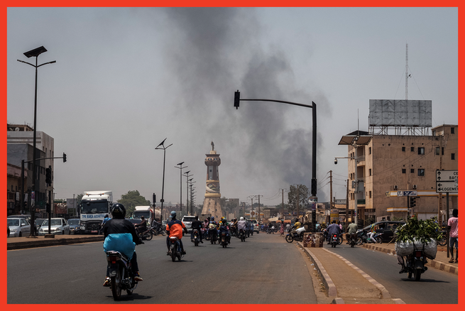 color photo of wide street with numerous mopeds, with traffic circle and statue and black smoke rising in distance