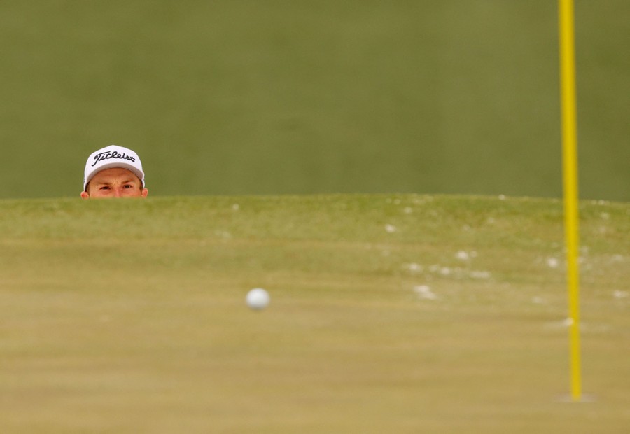 The head of a golfer behind a hill as a golf ball rolls toward a pin