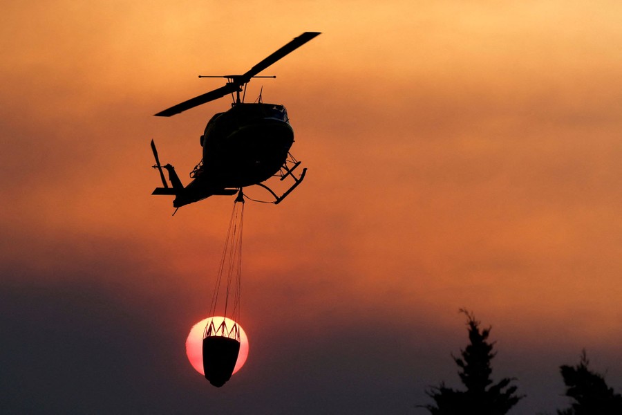 A firefighting helicopter carrying a water bucket flies in front of the sun.