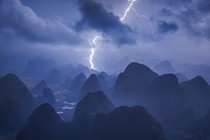 An aerial image of clusters of steep mountains seen in stark contrast beneath a cloudy night sky and a single bolt of lightning
