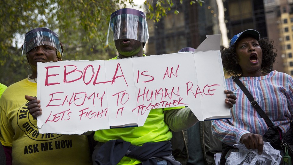 Three people at a march. Two of them are wearing face shields. One holds a sign that reads, "Ebola is an enemy to human race. Let's fight it together."