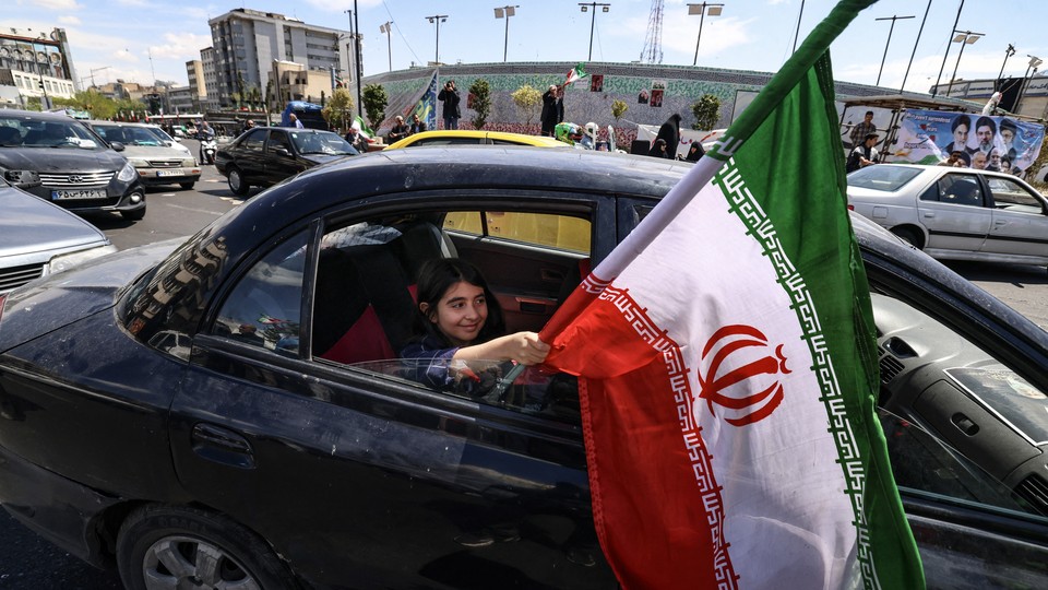 Photograph of a young girl sitting in the backseat of a car and holding an Iranian flag out of the rolled down window