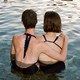 two people in bathing suits in lake, facing away from the camera, with an arm around each other