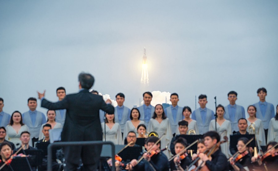 An orchestra and chorus perform in front of a distant launching rocket.