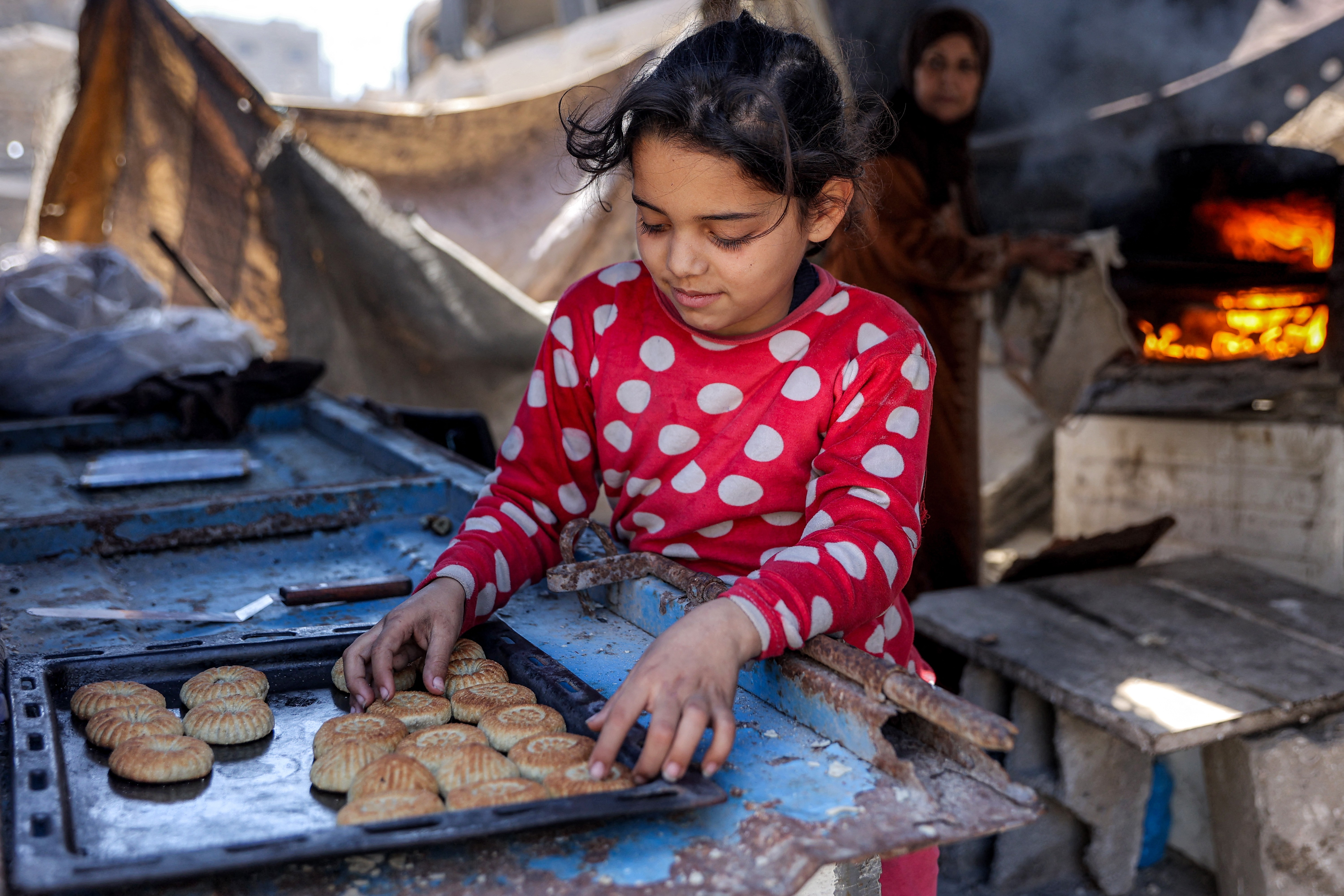 A girl sorts biscuits into a tray while a person behind her tends to an oven.