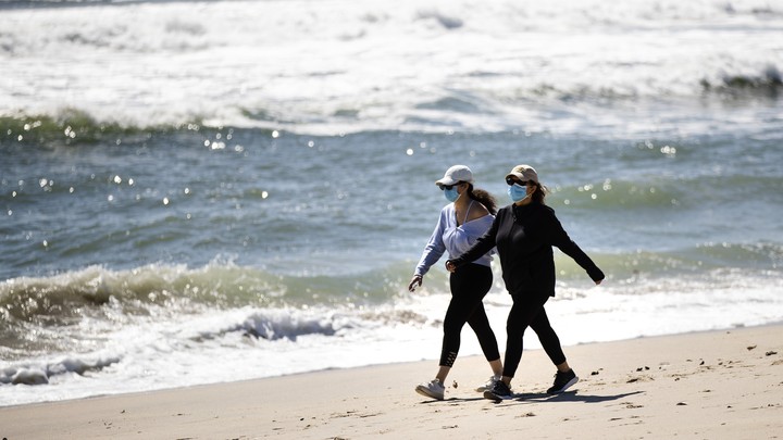 Two women wearing face masks walk on a beach
