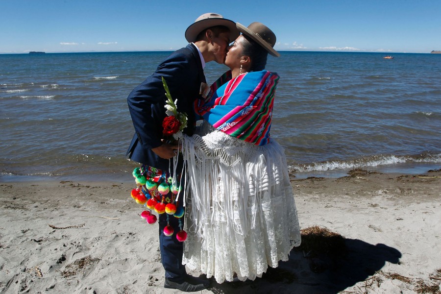 A newlywed couple kiss each other while standing on the shore of a lake.