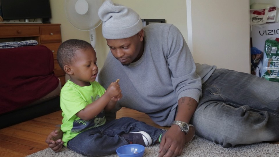 A father sits with his son on the carpet and looks at him lovingly while the child stares at a snack.