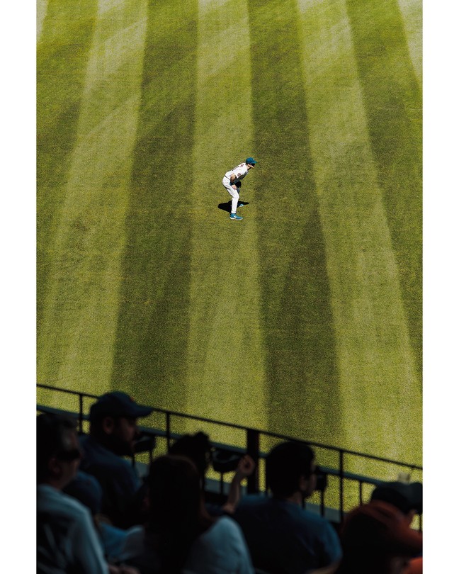 A photograph of a baseball outfielder on green field with fans silhouetted in the foreground.
