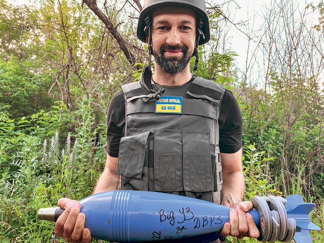 photo of smiling man in flak vest and helmet holding large blue torpedo-shaped bomb